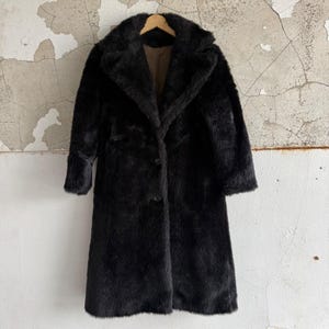 May include: A long, black faux fur coat featuring a wide notched collar, long sleeves, and a front button closure. The interior lining is a solid brown fabric. The coat is displayed on a wooden hanger, showcasing a thick, textured pile.