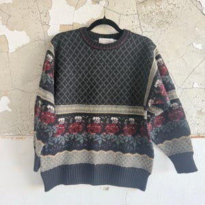 May include: A black sweater with a diamond pattern and floral accents. The sweater features red roses and green leaves, with a ribbed hem and cuffs. The sweater is hanging on a black hanger.