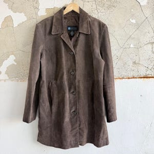 May include: A brown suede coat with a collared neckline and button-down closure. The coat has long sleeves and a tailored fit. The coat is hanging on a wooden hanger against a distressed wall.