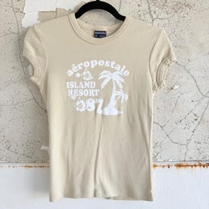 May include: A beige short-sleeved t-shirt with a white graphic print on the chest. The design features the text 'aeropostale ISLAND RESORT 87' alongside illustrations of a palm tree and hibiscus flowers. The shirt has a crew neckline.