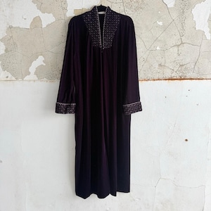 Puede incluir: Una bata larga de terciopelo morado oscuro con mangas largas, colgada contra una pared blanca desgastada. La bata presenta un ribete bordado decorativo alrededor del cuello y los puños. Es una prenda vintage.