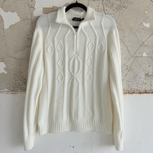 May include: A cream-coloured knitted sweater with a half-zip closure and a ribbed collar. The sweater features a cable-knit design down the centre and diamond-shaped patterns. The long sleeves have ribbed cuffs, and it hangs on a black hanger.