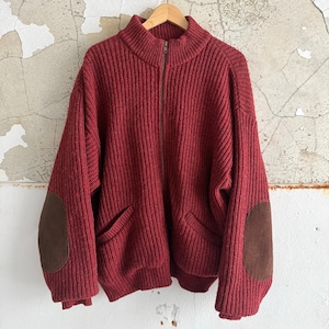 May include: A burgundy-colored, ribbed-knit zip-up sweater with a high collar and two front pockets. The sweater features brown suede elbow patches. The garment is hanging on a wooden hanger against a distressed wall.