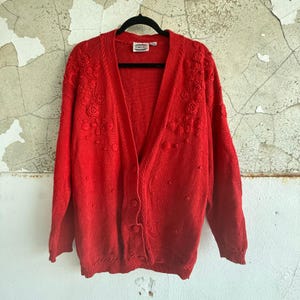 Puede incluir: Un cárdigan rojo brillante con cuello en V y cierre de botones. El suéter presenta bordados florales en los hombros y pequeños puntos en relieve en todo el tejido. Cuelga de una percha negra.