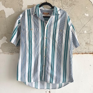 May include: Short-sleeved button-down shirt with vertical stripes in white, teal, and navy blue. The shirt has two chest pockets and a collar. The brand name "RUSTLER" is visible on the label. The shirt is hanging on a black hanger.