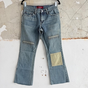 May include: Light blue denim jeans with a faded wash and a slight flare at the hem. The jeans feature patches on the knees and lower leg. The jeans are hanging on a black hanger.