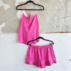 May include: A bright pink pajama set, including a camisole top and matching shorts, hangs on black hangers against a textured wall. The camisole has thin straps and a racerback design. The shorts have a white trim.