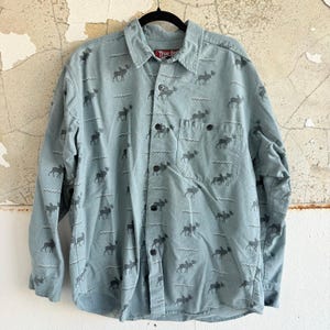 May include: A pale blue button-down shirt featuring a pattern of grey moose and wavy lines. The shirt has a chest pocket and the brand name "True Grit" is visible on the collar.