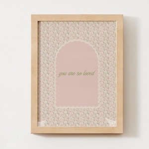 May include: A framed print with a light wood frame. The print features a pink background with the words "you are so loved" in green script. The print has a floral pattern and a decorative arch.