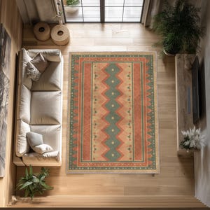 Pode incluir: Vista aérea de um tapete retangular com um design geométrico. O tapete apresenta um padrão central de diamantes e ziguezagues em tons de verde, pêssego e bege, emoldurado por uma borda de cores semelhantes. O tapete está sobre um chão de madeira.