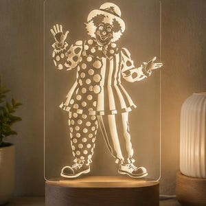 Puede incluir: Una l&aacute;mpara LED rectangular de acr&iacute;lico con un dise&ntilde;o de payaso grabado. El payaso lleva un sombrero, un cuello con volantes y un traje dividido entre lunares y rayas verticales. El panel de acr&iacute;lico transparente se asienta sobre una base de madera.