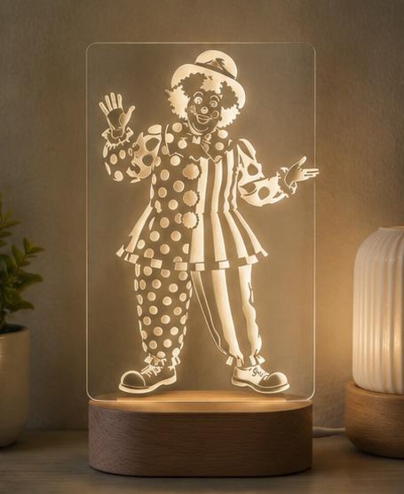 Puede incluir: Una l&aacute;mpara LED rectangular de acr&iacute;lico con un dise&ntilde;o de payaso grabado. El payaso lleva un sombrero, un cuello con volantes y un traje dividido entre lunares y rayas verticales. El panel de acr&iacute;lico transparente se asienta sobre una base de madera.