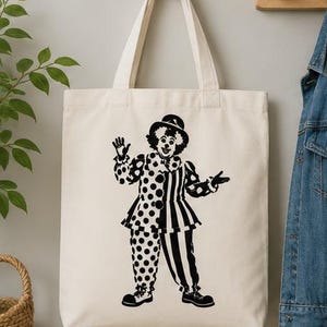Puede incluir: Un bolso de tela color crema con un estampado gr&aacute;fico negro de un payaso que lleva un sombrero, un cuello con volantes y un disfraz dividido entre lunares y rayas verticales. El payaso aparece de pie con una mano levantada saludando.