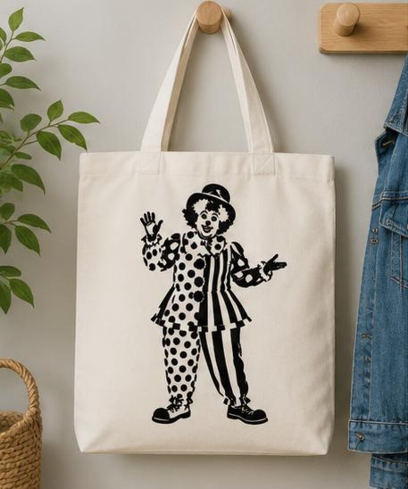Puede incluir: Un bolso de tela color crema con un estampado gr&aacute;fico negro de un payaso que lleva un sombrero, un cuello con volantes y un disfraz dividido entre lunares y rayas verticales. El payaso aparece de pie con una mano levantada saludando.
