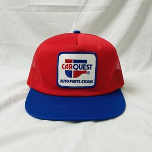 May include: A red and blue trucker hat with a mesh back. The hat features a rectangular patch with the text "CARQUEST AUTO PARTS STORES" in white and blue. The hat has a blue brim and a red crown.