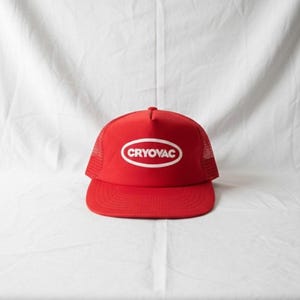 May include: A red trucker hat featuring a white oval logo with the text "CRYOVAC". The hat has a mesh back and a curved peak. The hat is displayed on a white background.