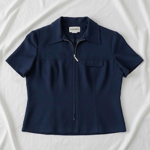 Puede incluir: Top azul marino de manga corta con cuello y cremallera completa. El top tiene un pequeño bolsillo en el lado derecho y la etiqueta "PLAZA SOUTH" es visible. La prenda está hecha de tela lisa.