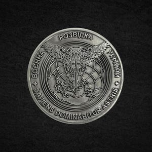 May include: A silver coin with intricate detailing. The coin features an owl with outstretched wings above a globe, surrounded by text in Ukrainian and Latin. The words "Sapien Dominabitur Astris" are visible.