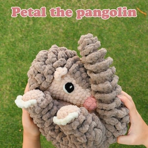 May include: A crocheted pangolin plush toy with a light beige face, a pink nose, and a textured brown body. The text on the image reads: Low sew PDF pattern. Only the head is sewn on! Petal the pangolin. The plush has white accents on its paws.