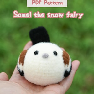 Puede incluir: Una figura de pájaro de fieltro con un cuerpo redondo blanco, parches de alas marrones, plumas de cola negras y ojos negros pequeños. El texto sobre el pájaro dice PDF Pattern y Somei the snow fairy en fuente rosa claro sobre fondo blanco.