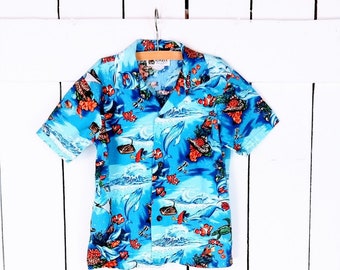 Puede incluir: Una camisa azul de manga corta con estampado tropical que incluye peces, corales y conchas. La camisa tiene un cierre de botones en la parte delantera y un cuello.