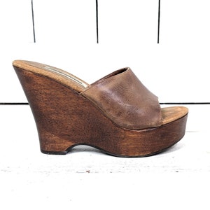 May include: Brown leather wedge sandals with a wooden platform sole. The sandals have a peep toe and a slip-on design.