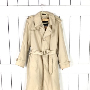 May include: A tan trench coat with a belted waist and button closures. The coat features a classic design with a collar, epaulets, and adjustable cuffs. It is hanging on a white hanger against a white wooden background.