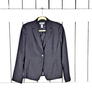 Può includere: Un blazer nero con una chiusura a un solo bottone. Il blazer è appeso a un appendiabiti di legno su uno sfondo di pannello di legno bianco.