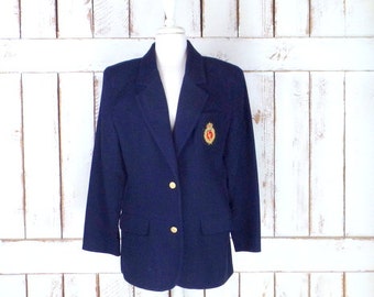 Könnte beinhalten: Ein marineblauer Blazer mit goldenen Knöpfen und einem gestickten Wappen in Gold und Rot auf der linken Seite.