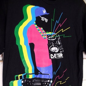Puede incluir: Una camiseta negra con un estampado gráfico colorido de una persona que lleva una gorra de béisbol azul con las letras "LA" en ella, una camisa rosa, pantalones azules y un collar verde y amarillo. La persona sostiene una gran radiocasete con la palabra "DETER" impresa en ella. El gráfico está rodeado de rayos coloridos.
