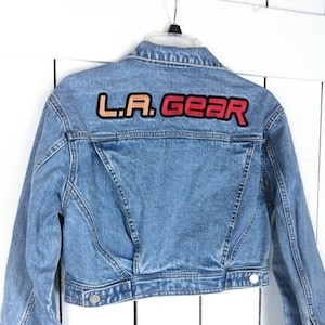 Könnte beinhalten: Eine hellblaue Jeansjacke mit verkürzter Passform. Die Jacke hat einen Aufnäher auf der Rückseite, auf dem "L.A. GEAR" in roten, orangen und gelben Buchstaben steht.