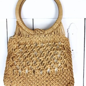 Op de afbeelding: Een beige gehaakte tote bag met een houten handvat. De tas is gemaakt van dik, geweven touw en heeft een losse, open weving.