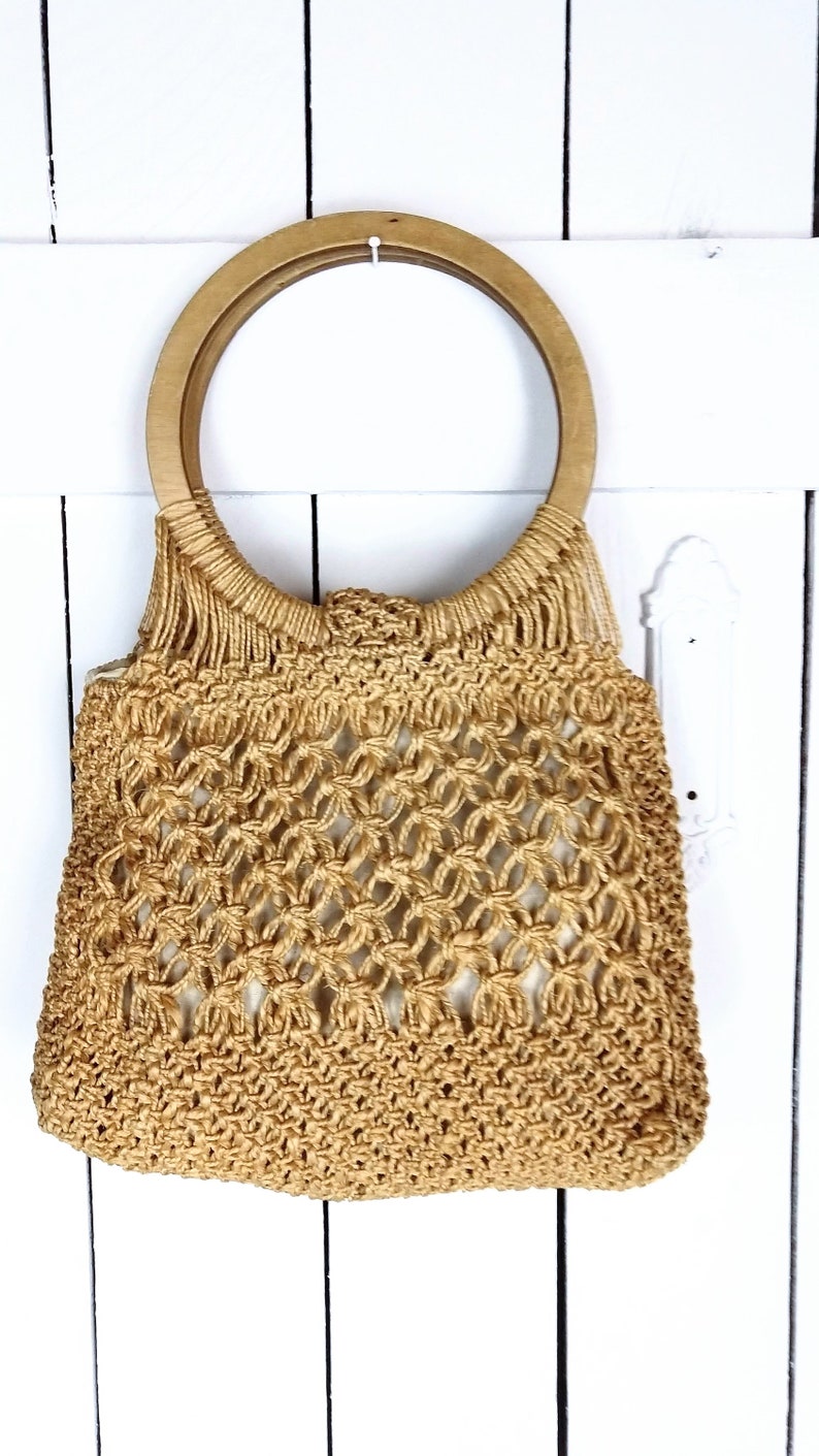 Op de afbeelding: Een beige gehaakte tote bag met een houten handvat. De tas is gemaakt van dik, geweven touw en heeft een losse, open weving.