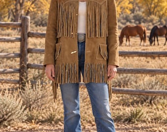 May include: A tan suede jacket featuring long fringe detailing on the chest and hem, a collared neckline, and toggle closures. The jacket is worn with a white top, blue denim jeans with a large belt buckle, and a tan cowboy hat with a dark band.