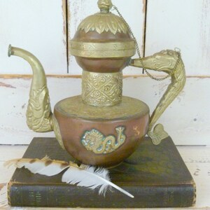 May include: A copper and brass teapot with a dragon design and a chain handle. The teapot is sitting on a dark brown book with a white feather on top.