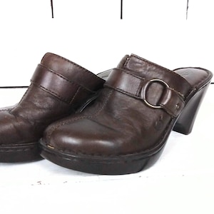 May include: A pair of brown leather heeled clogs with a buckle strap and a metal ring. The clogs have a chunky heel and a rounded toe.