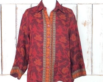 May include: A vintage button-down shirt in shades of red and orange, featuring a floral pattern. The shirt has a collared neckline, long sleeves, and a decorative orange stripe down the center. The shirt is displayed on a white mannequin.