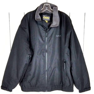 Puede incluir: Chaqueta negra de Cabela's con cuello de forro polar gris y cremallera completa. La chaqueta tiene un pequeño bolsillo en el pecho y el logotipo de Cabela's es visible en el pecho y el cuello. La chaqueta está colgada en una percha.
