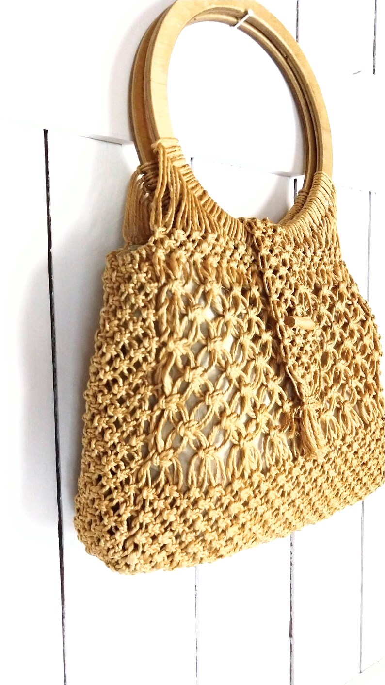 Op de afbeelding: Een beige gehaakte tote bag met een houten handvat. De tas heeft een geweven patroon en is gemaakt van natuurlijke vezels.