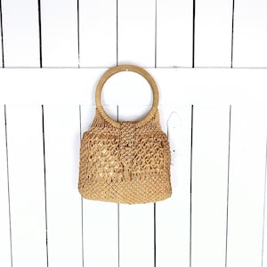 Op de afbeelding: Een beige geweven stro tas met een houten handvat. De tas heeft een geweven patroon en hangt aan een witte houten muur.
