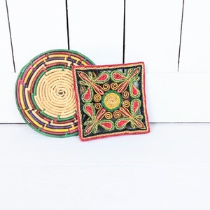 May include: Two woven straw trivets, one round with green, red and yellow stripes, and one square with a red, yellow and green floral design.