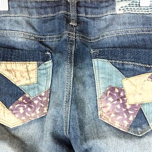 Könnte beinhalten: Eine blaue Jeans mit Patchwork-Details auf den Gesäßtaschen. Die Patches sind aus verschiedenen Stoffen in Blautönen, Braun und Lila.