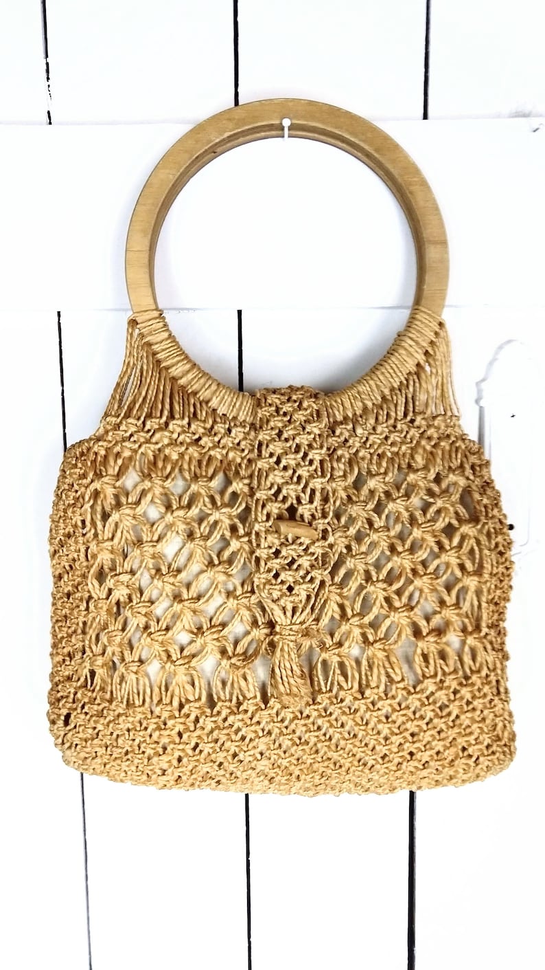 Op de afbeelding: Een beige gehaakte tote bag met een ronde houten handvat. De tas is gemaakt van geweven touwmateriaal.
