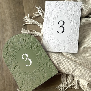 May include: A sage green arched table number card featuring a white embossed floral border and the number 3 in white, alongside a rectangular white table number card with a matching embossed floral border and the number 3 in black.