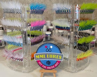 Può includere: Un'esposizione di esche da pesca colorate in espositori trasparenti in acrilico. Le esche presentano vari colori, tra cui bianco, blu, rosa e verde. Un piccolo cartello con la scritta "MML LURES" è di fronte.