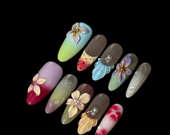 May include: A collection of ten long, almond-shaped artificial nails. Each nail features a unique design, including 3D floral embellishments, pearl accents, and colorful gradients. The nails showcase a variety of colors, including pink, green, brown, and white.