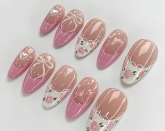 May include: A set of ten almond-shaped artificial nails. Each nail features a pink base with a nude-colored center. Designs include white bows, hearts, pearls, and floral patterns. The nails are arranged on a white surface.
