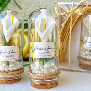 Puede incluir: Botellas de vidrio transparente con calas blancas, perlas y bases de corcho. Cada botella tiene una etiqueta con el texto "Alessia & Lorenzo" y "Thank you for coming". Las botellas están atadas con hilo.