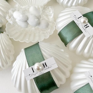 May include: White seashell-shaped containers, each adorned with a green ribbon and a pearl. Small tags with text and pearls are attached. Some shells are open, showing white candies inside.
