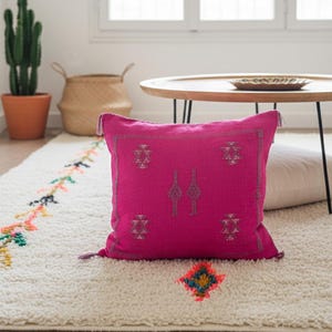 Op de afbeelding: Een felroze decoratief kussen met geborduurde geometrische patronen en kwastjes. Het kussen ligt op een crèmekleurig tapijt met kleurrijke geborduurde details. Een cactus en een ronde houten tafel staan op de achtergrond.